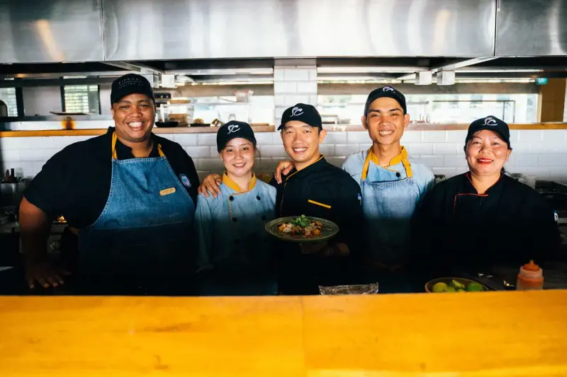 Smiling restaurant kitchen team in caps and aprons behind the counter holding a plated dish