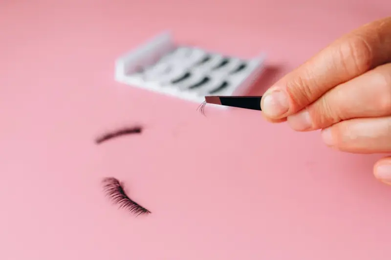 Tweezers holding false eyelashes over a pink background