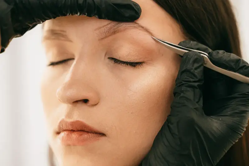 Gloved beautician shaping a client's eyebrows with tweezers