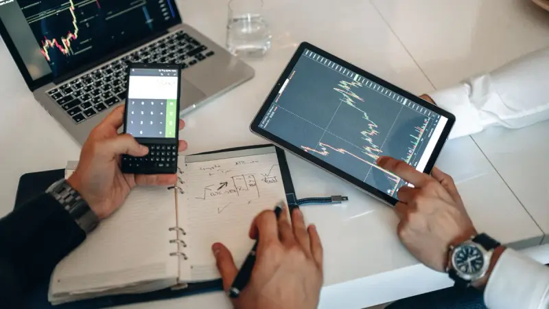 Hands holding a phone and tablet showing market charts over a notebook