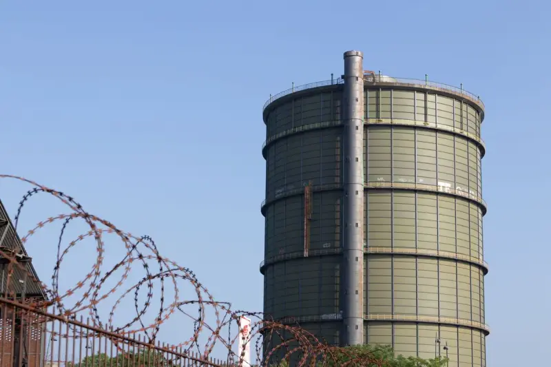 Tall cylindrical green gasometer behind a fence topped with razor wire