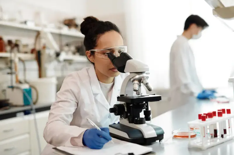 Woman scientist taking notes at a microscope with sample tubes nearby