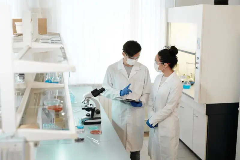 Two scientists in lab coats discussing results beside a microscope in a bright lab