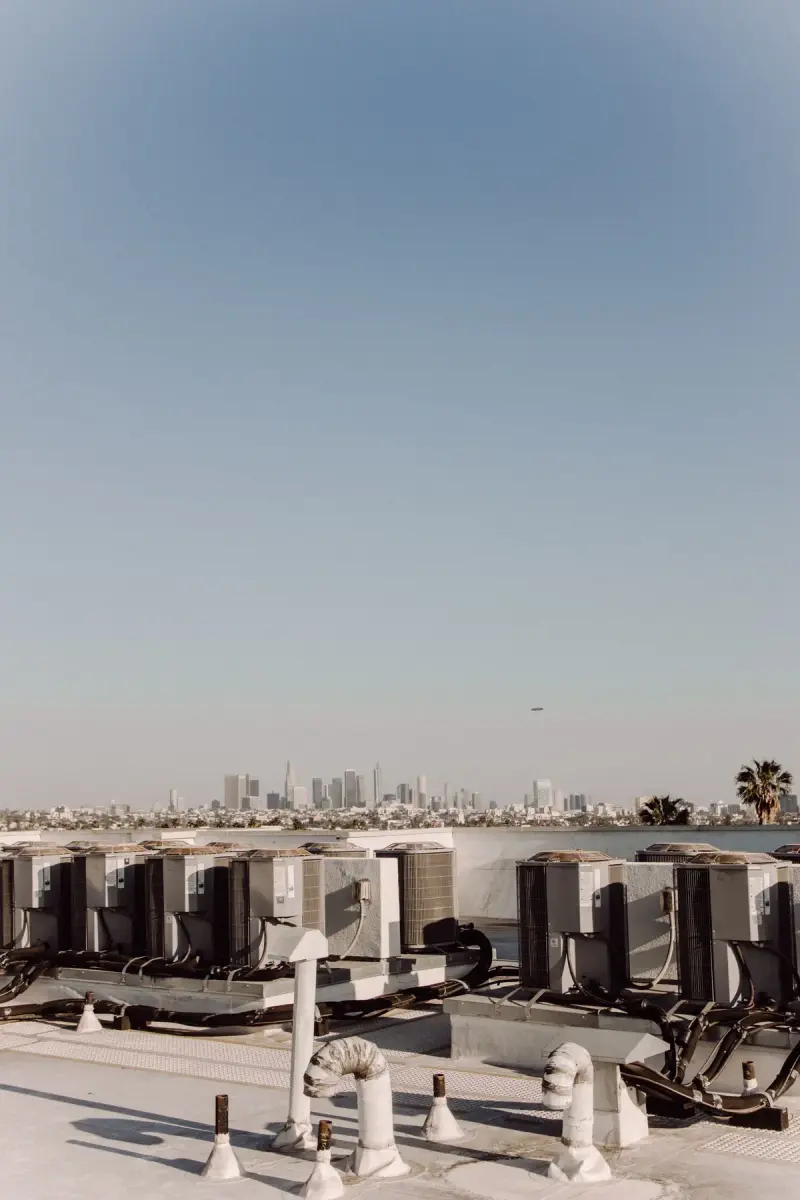 Rows of HVAC condenser units on a flat building roof with a city skyline behind