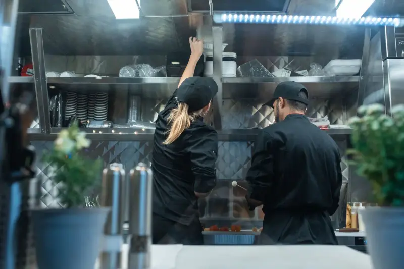 Woman and man in black chef uniforms reaching for supplies on stainless kitchen shelves