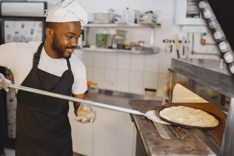 Smiling cook in a white hat sliding a pizza from an oven with a metal peel