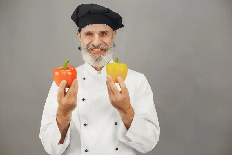 Smiling chef in a white jacket and black toque holding a red and a yellow bell pepper