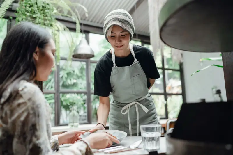 Server in a gray apron bringing a dish to a guest in a bright cafe with plants