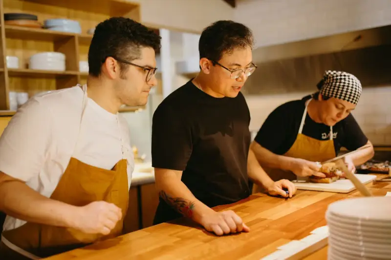 Three cooks in aprons working side by side at a wooden counter in a restaurant kitchen
