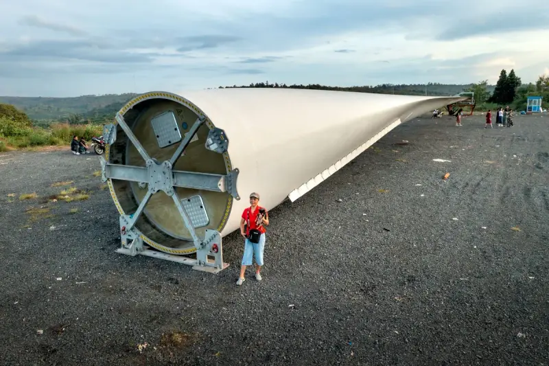 Full-length wind turbine blade lying on gravel, with a person beside the root for scale