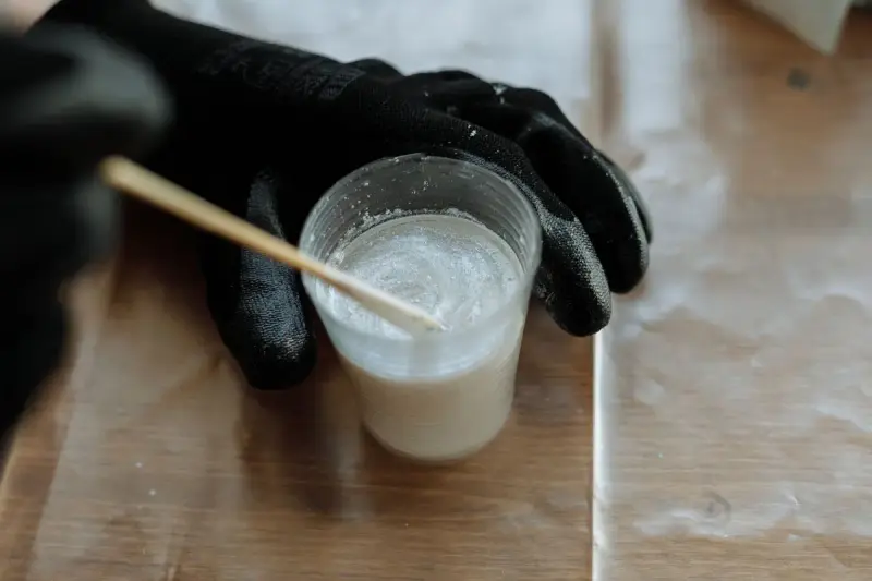 Black-gloved hand stirring milky resin in a clear cup with a wooden stick