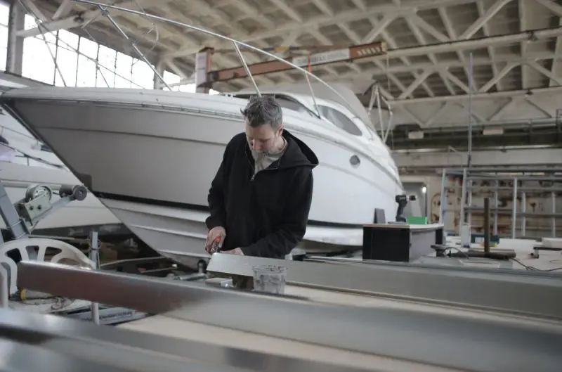 Bearded worker fitting a part at a bench beside a white boat hull in a large shed