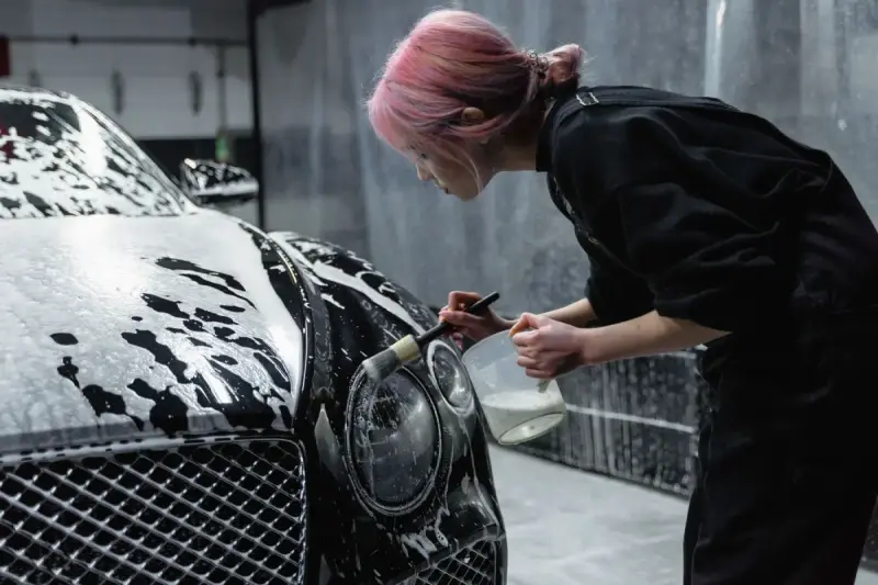 Woman cleaning a black car headlight with a detailing brush