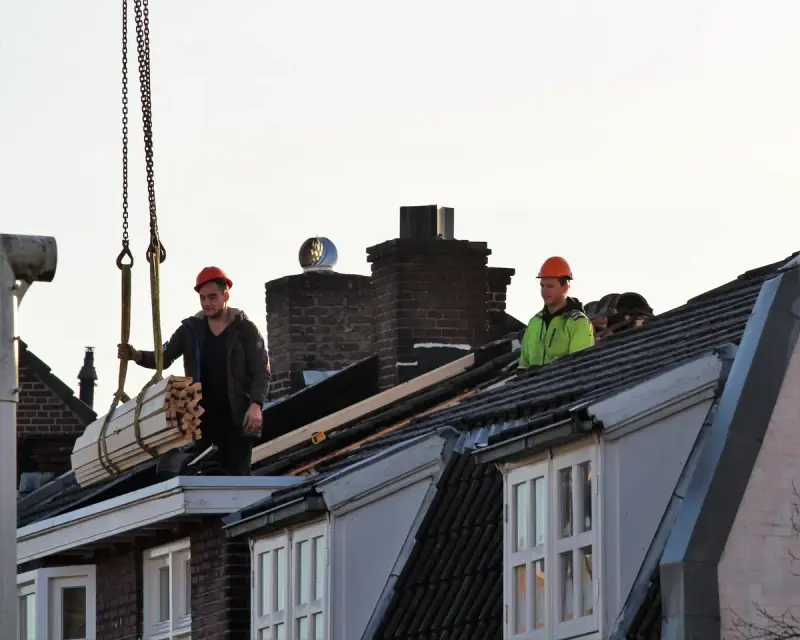Roofers in hard hats guiding a crane-lifted bundle onto a row of townhouse roofs