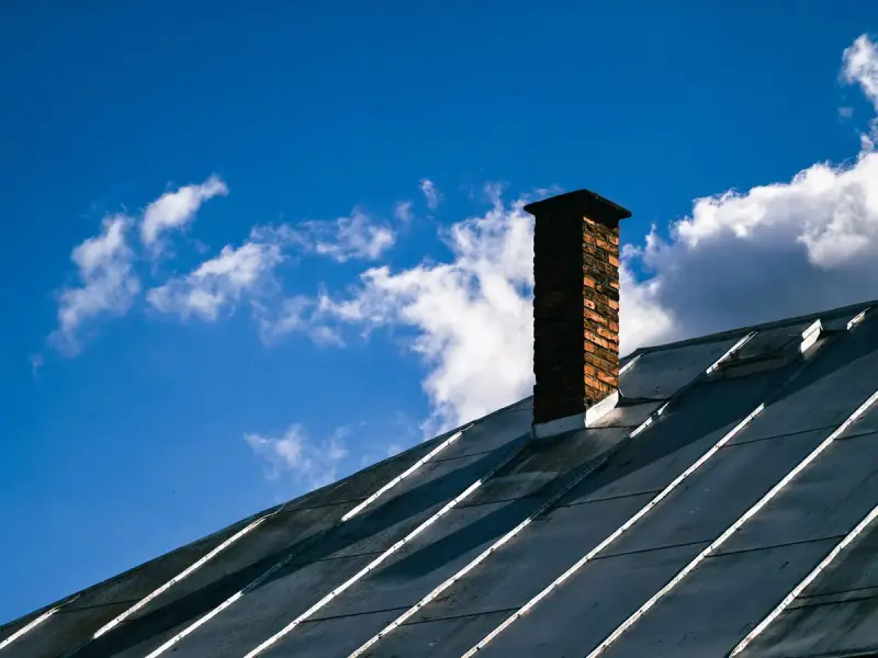 Dark standing-seam metal roof with a brick chimney against a bright blue sky
