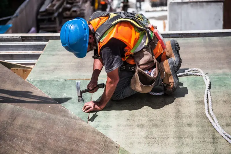 Roofer in a blue hard hat and harness hammering on a low-slope roof deck