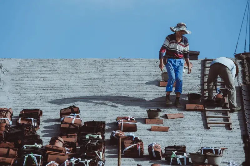 Two workers laying stacked clay roof tiles on a sunlit roof under a blue sky