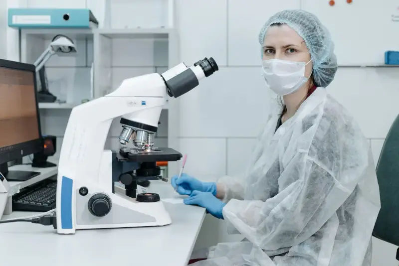 Masked lab scientist in protective gown seated at a microscope beside a computer
