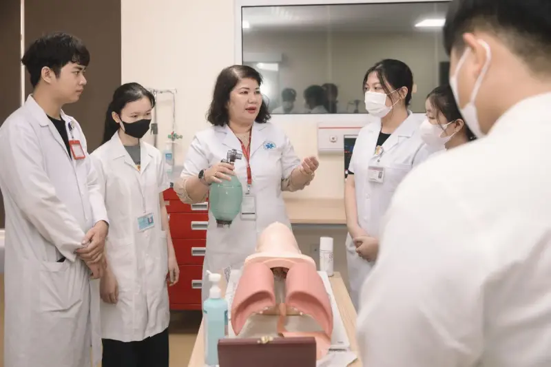Instructor holding a resuscitation bag beside an airway training model while trainees in white coats watch