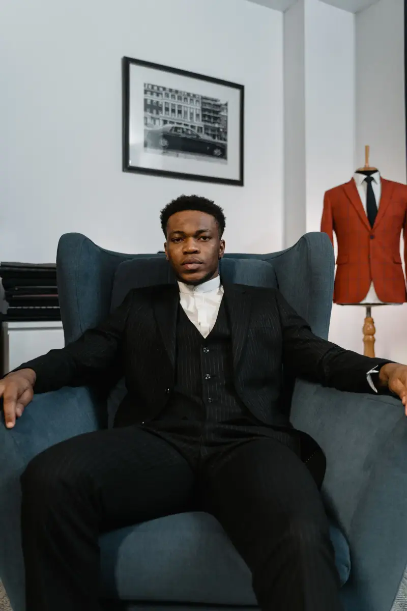 Man in a black three-piece suit seated in a blue wingback chair beside a red jacket form