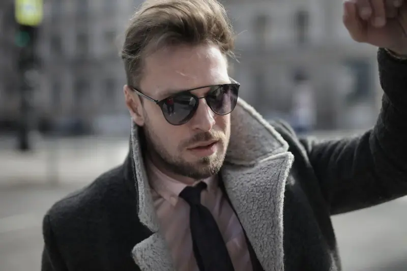 Man in sunglasses, a shawl-collar overcoat and knit tie adjusting his collar on a city street