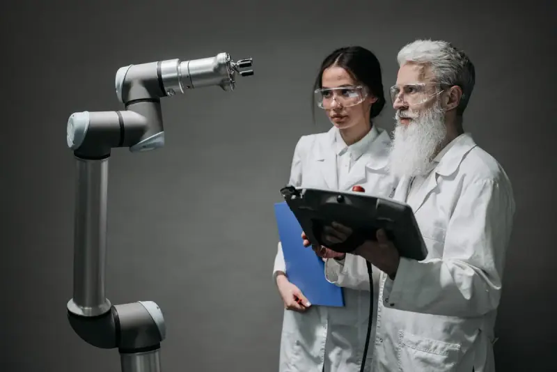 Two researchers in lab coats and safety glasses testing a robotic arm with a tablet