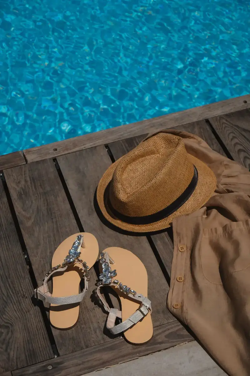 Straw fedora, beaded sandals and a linen shirt on a wooden deck beside a blue pool
