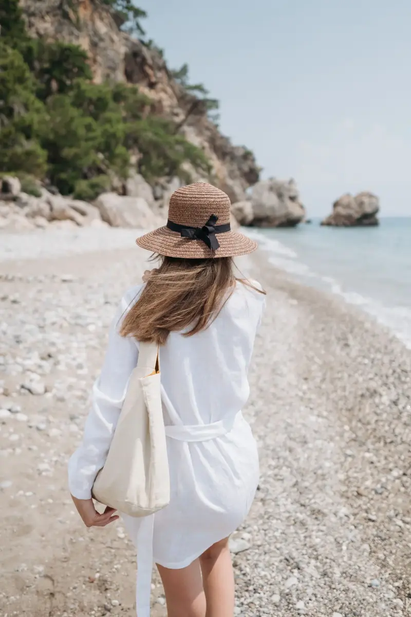 Woman in a white linen shirt dress and straw hat with a canvas tote walking a pebble beach