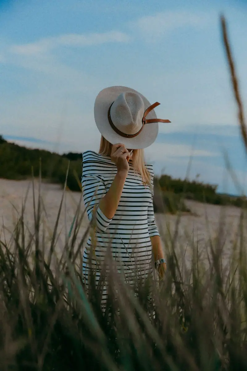 Woman in a striped knit dress holding a straw hat over her face in dune grass