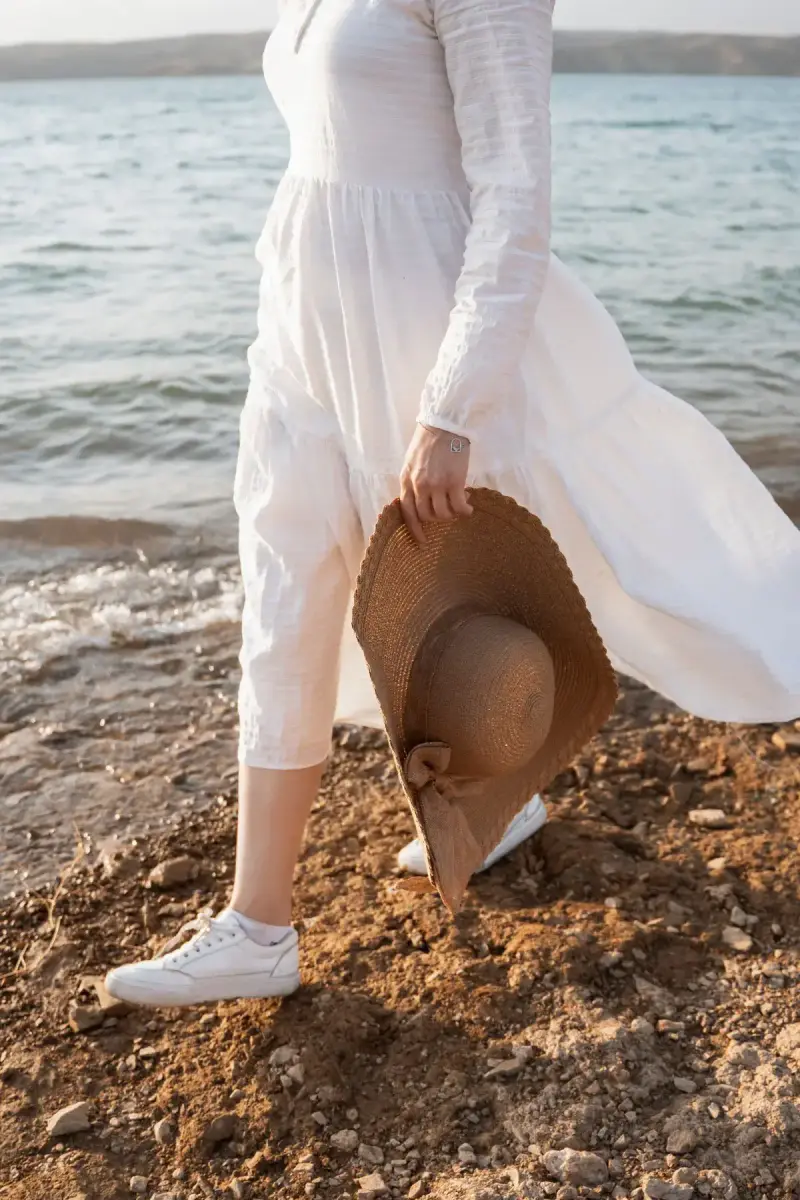 Woman in a white lace-sleeve maxi dress carrying a wide sun hat along the shoreline