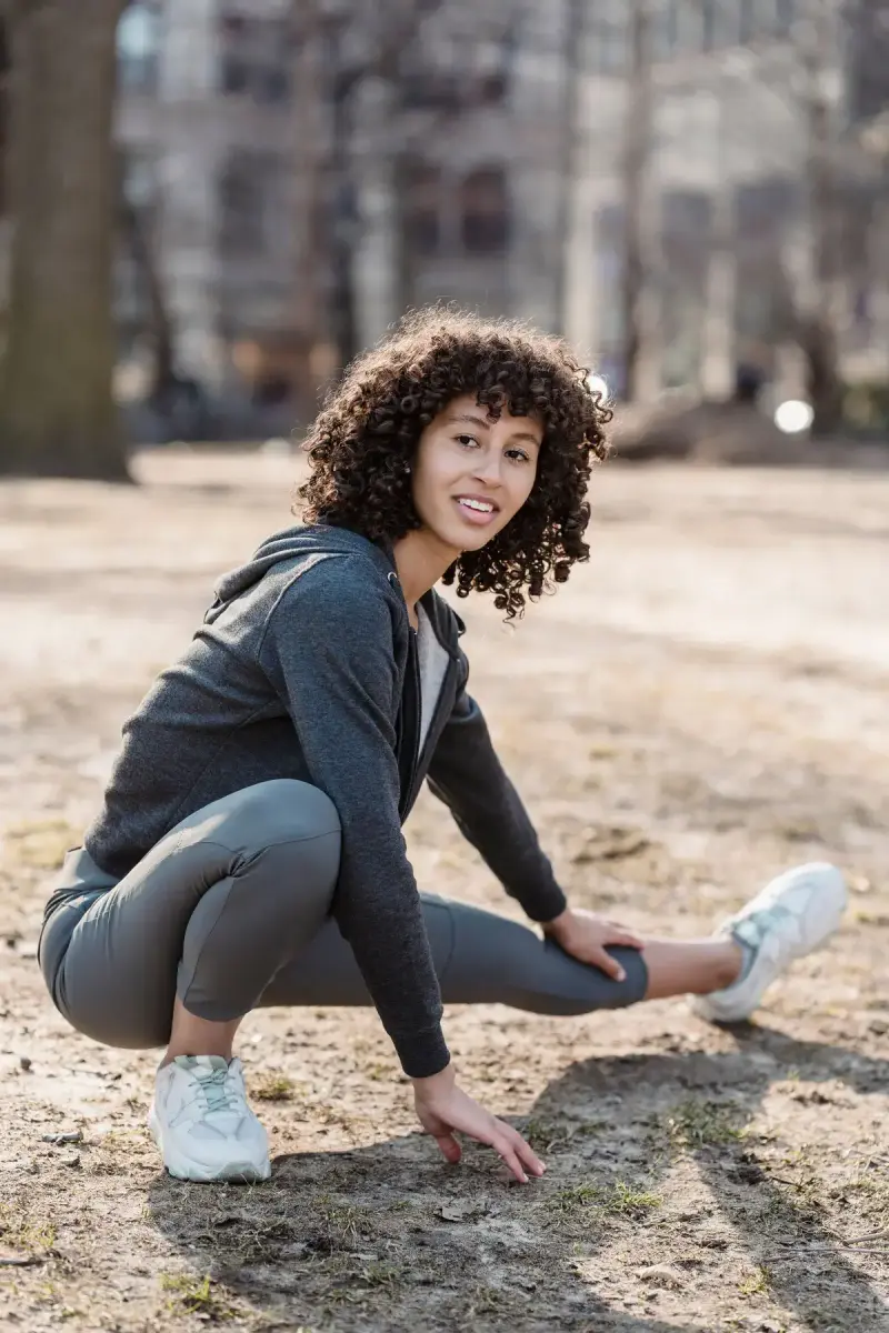 Smiling woman in a gray zip hoodie, leggings and white sneakers stretching in a park