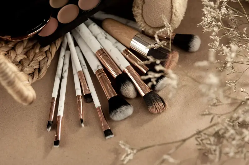 Rose-gold makeup brushes fanned out beside an eyeshadow palette on a beige surface