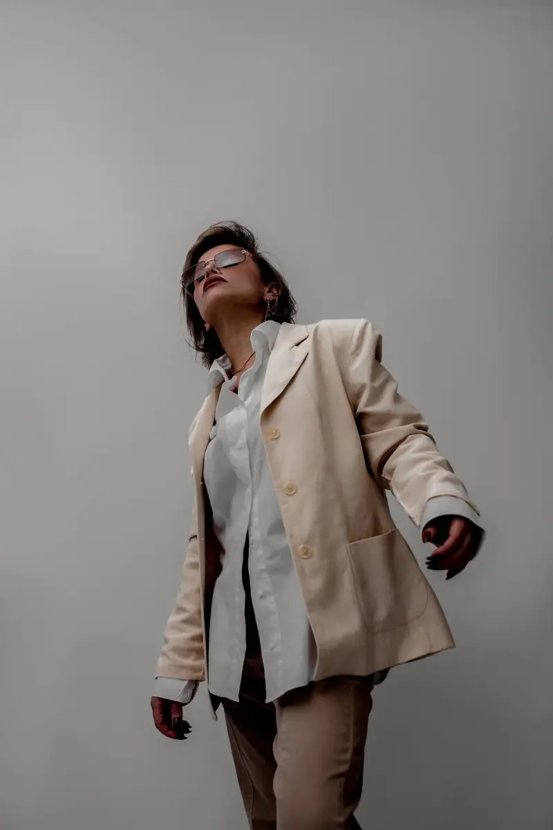 Woman in a sand-colored blazer, white shirt and tinted glasses posing against a gray studio wall