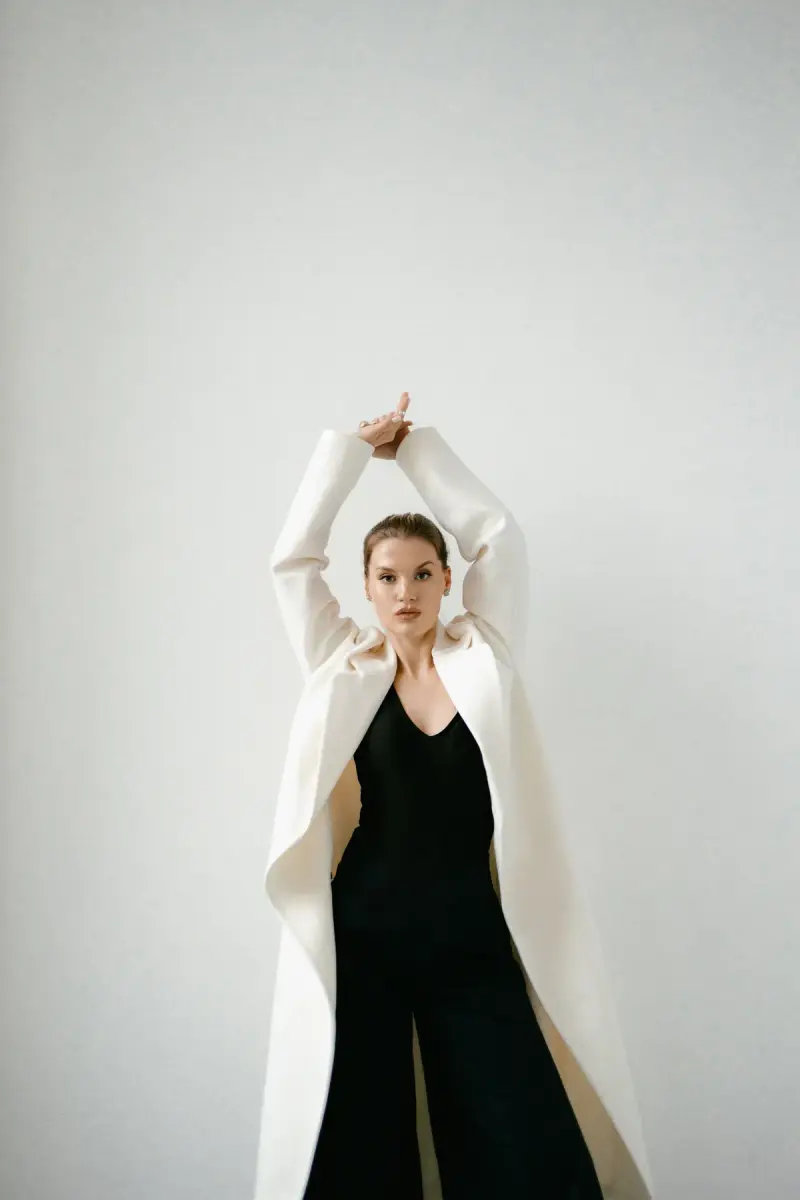 Woman in a long white coat over a black jumpsuit, arms raised against a white wall