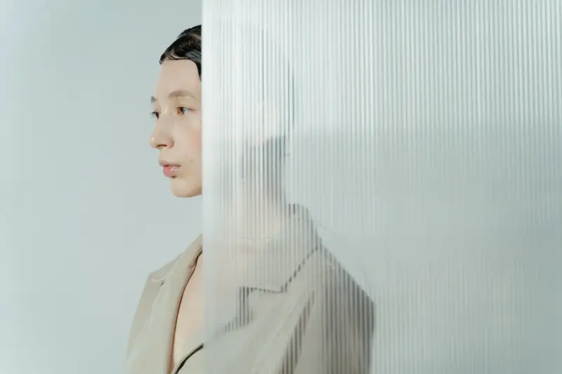 Woman in a beige coat seen half behind a ribbed glass panel in pale blue light