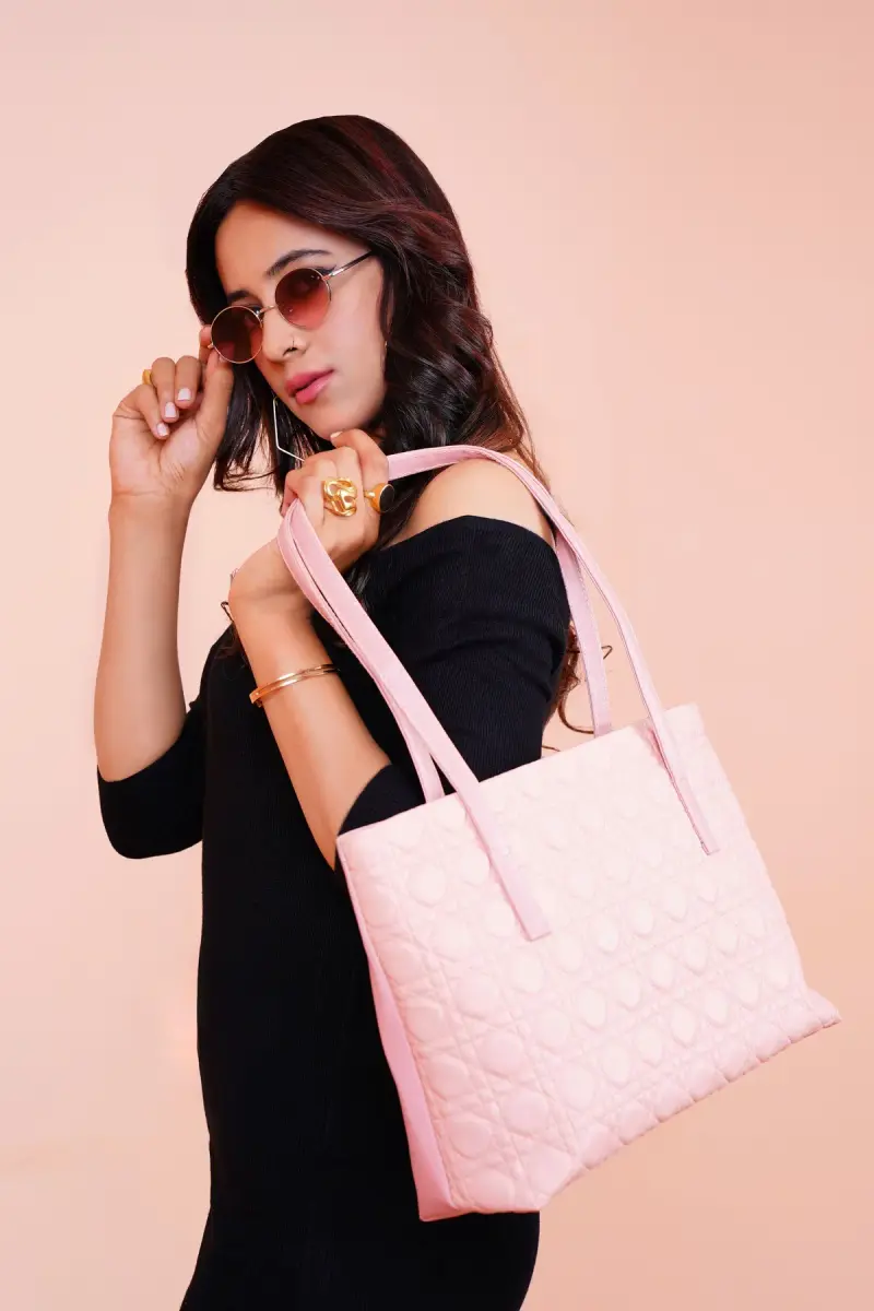 Woman in a black off-shoulder top and sunglasses with a quilted pink tote on a blush backdrop