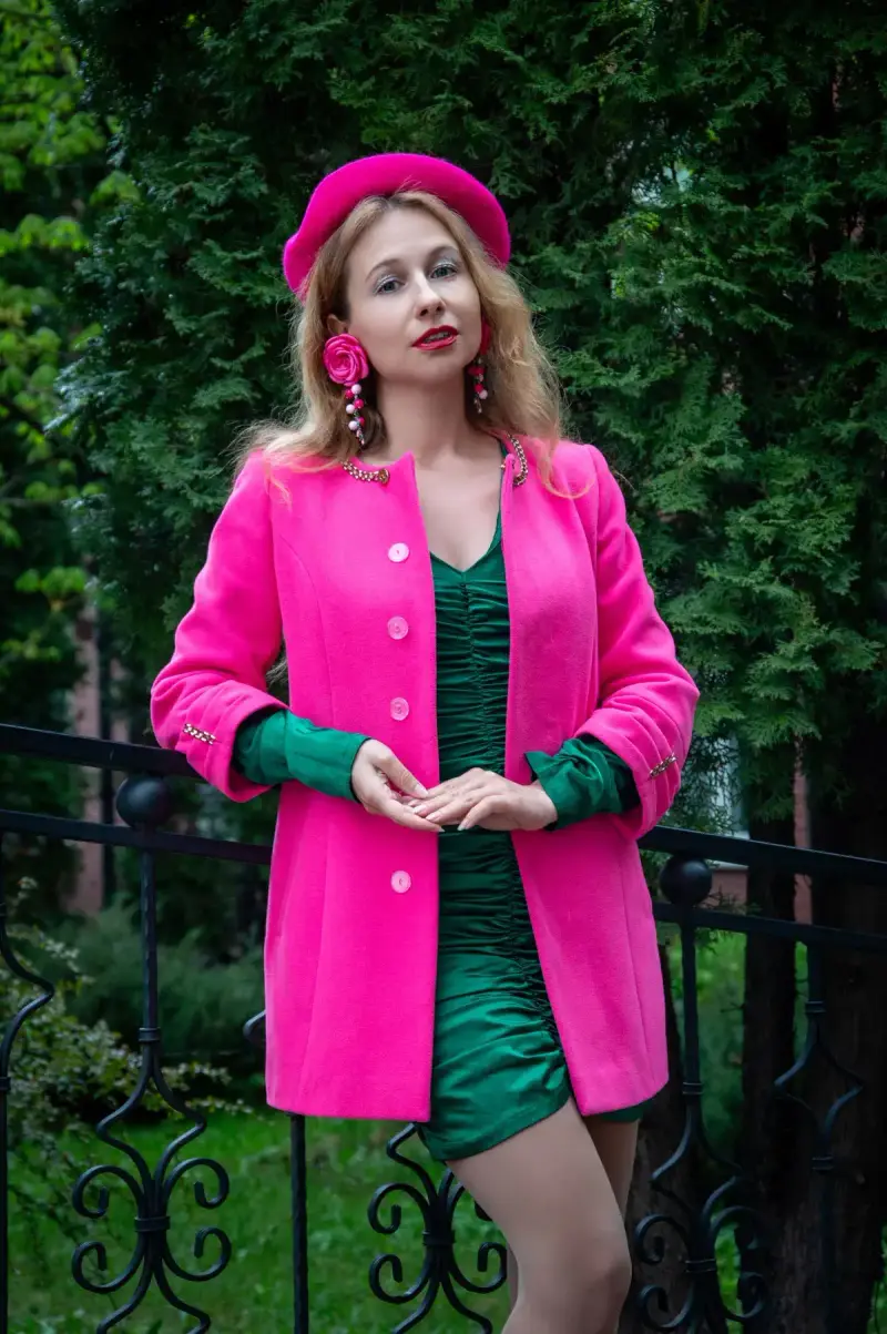 Woman in a hot pink coat and beret over an emerald dress by a garden gate