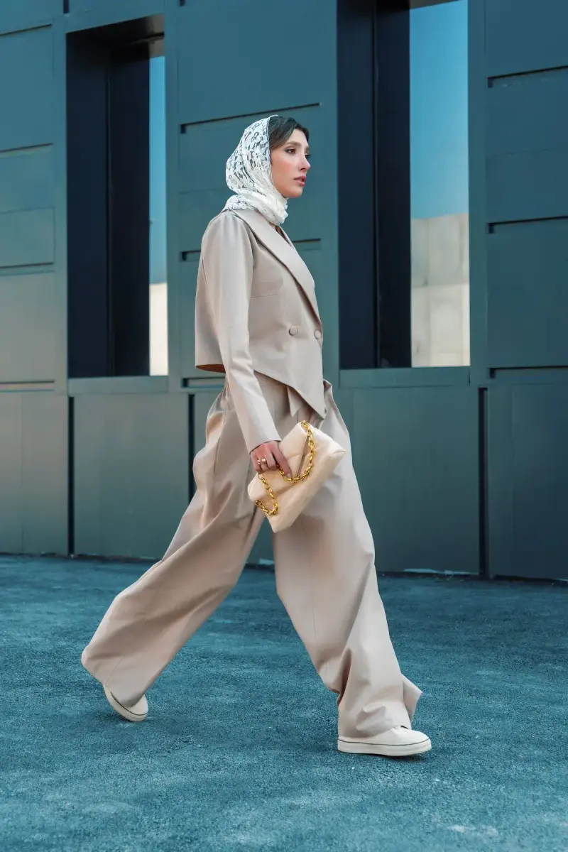 Woman in a beige oversized suit, silk headscarf and sneakers walking past teal walls