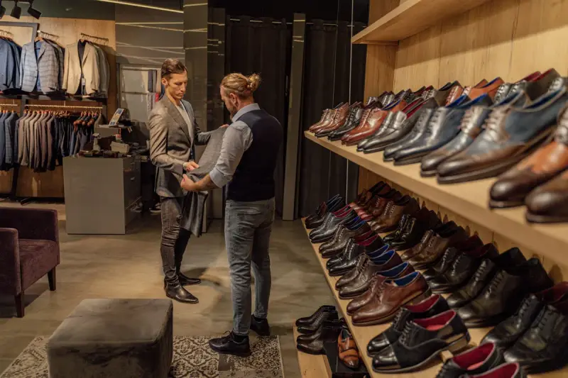 Men's clothing store with a long wooden shelf of leather dress shoes and a tailor fitting a jacket