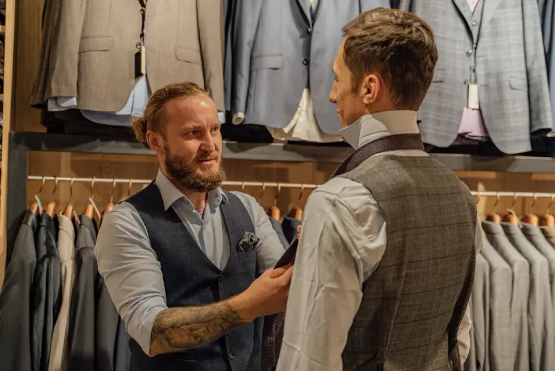 Clothier fitting a tie on a man in a tweed waistcoat in front of a wall of suits