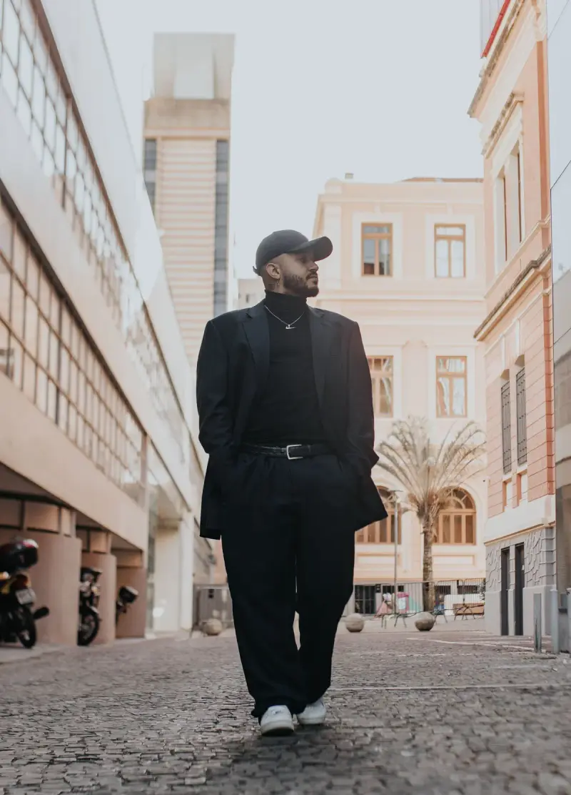 Man in a black suit, turtleneck and white sneakers walking a cobblestone street