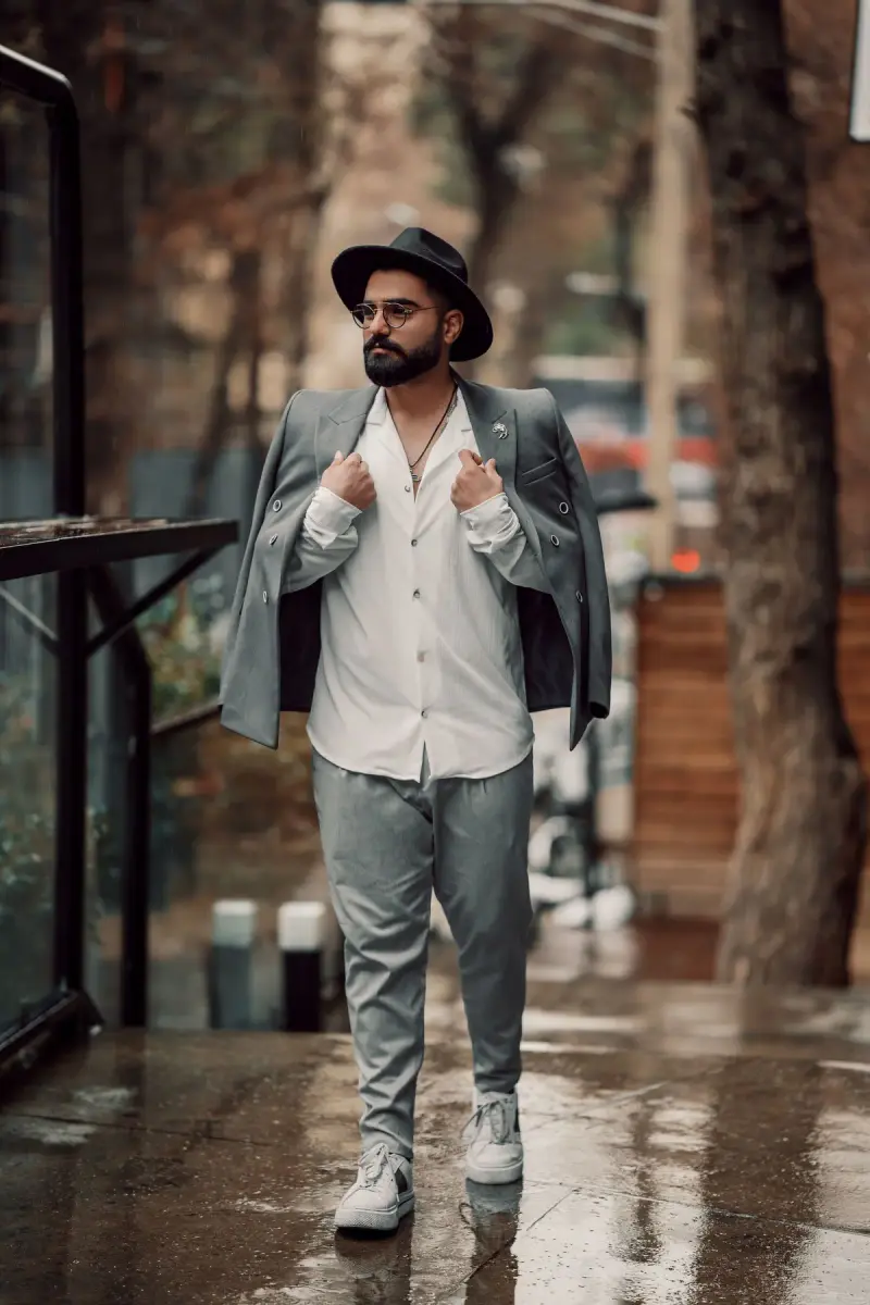 Man in a gray blazer, white shirt and felt hat walking a rain-wet path