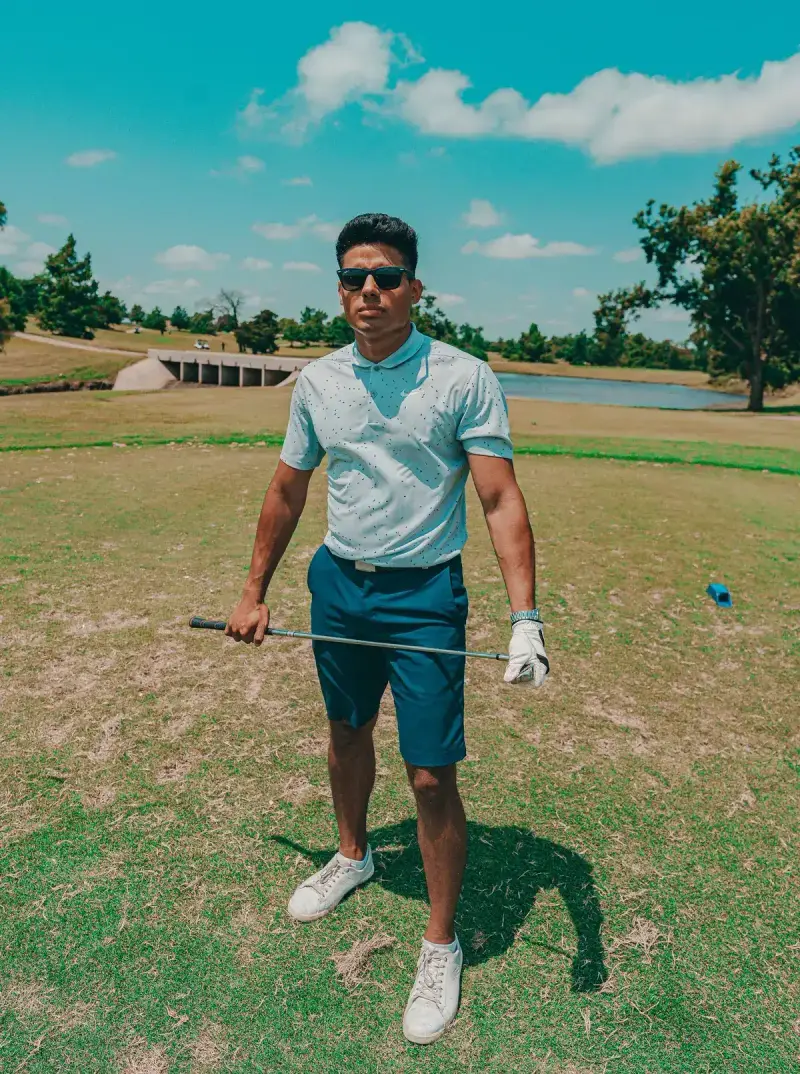 Man in sunglasses, a pale blue polo and navy shorts holding a club on a sunny golf course