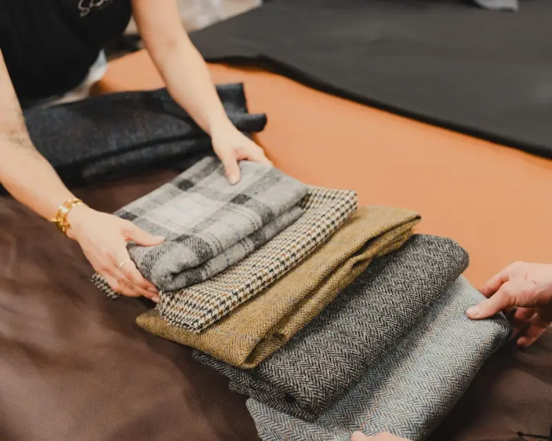 Hands arranging folded tweed and herringbone fabrics on a tailor's table