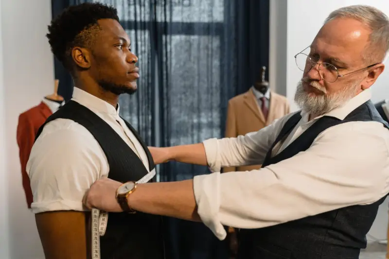 Grey-bearded tailor measuring a young man's chest with a tape in a workshop