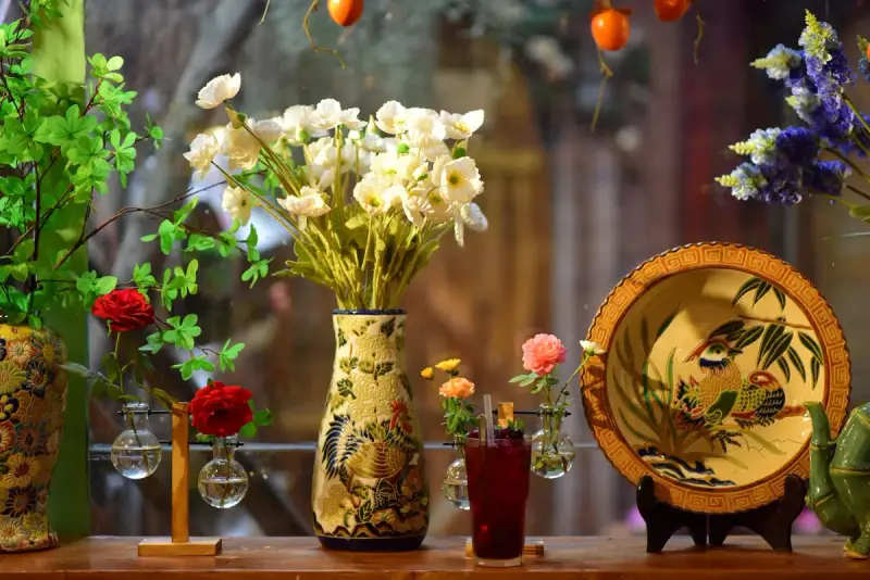 Painted ceramic vase of white flowers beside small glass bud vases and a decorative plate