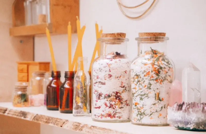 Glass jars of dried flower bath salts beside amber bottles, candles and a crystal on a shelf