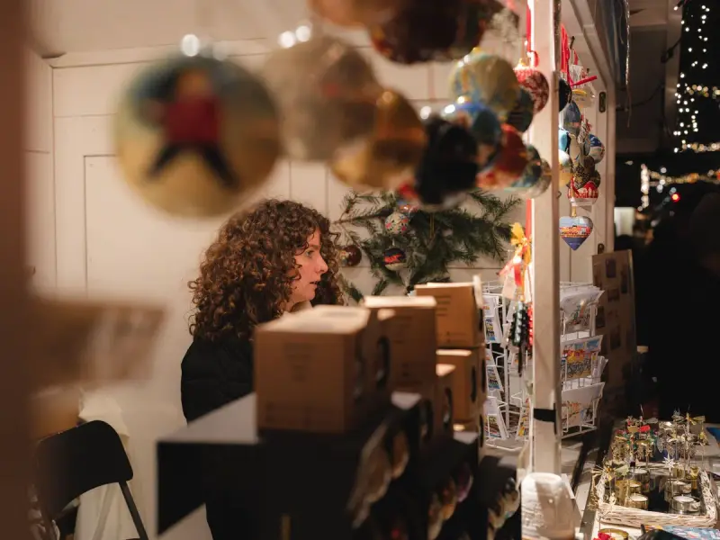 Woman tending a small holiday stall hung with painted ornaments and string lights