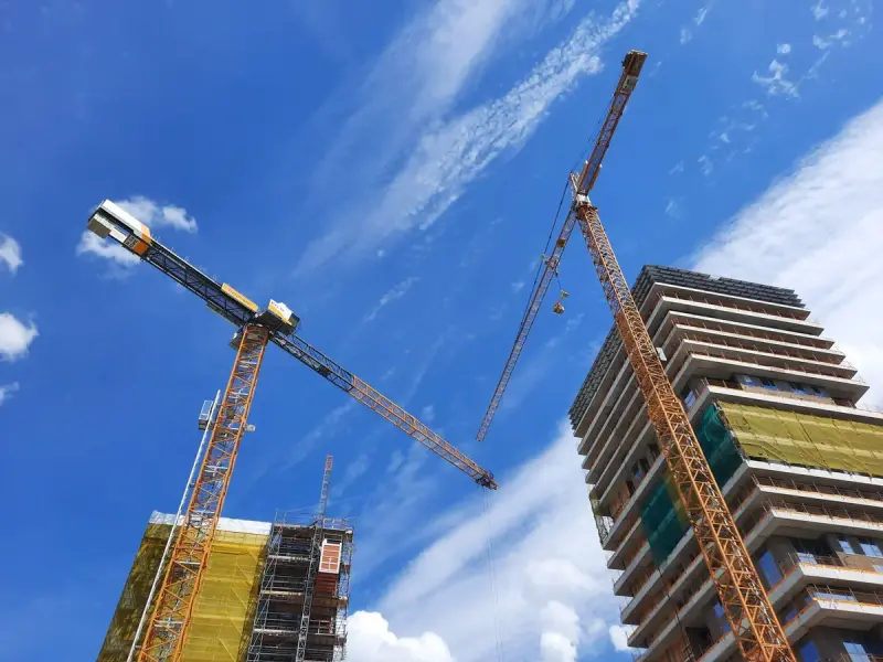 Low-angle view of tower cranes beside a building under construction against blue sky