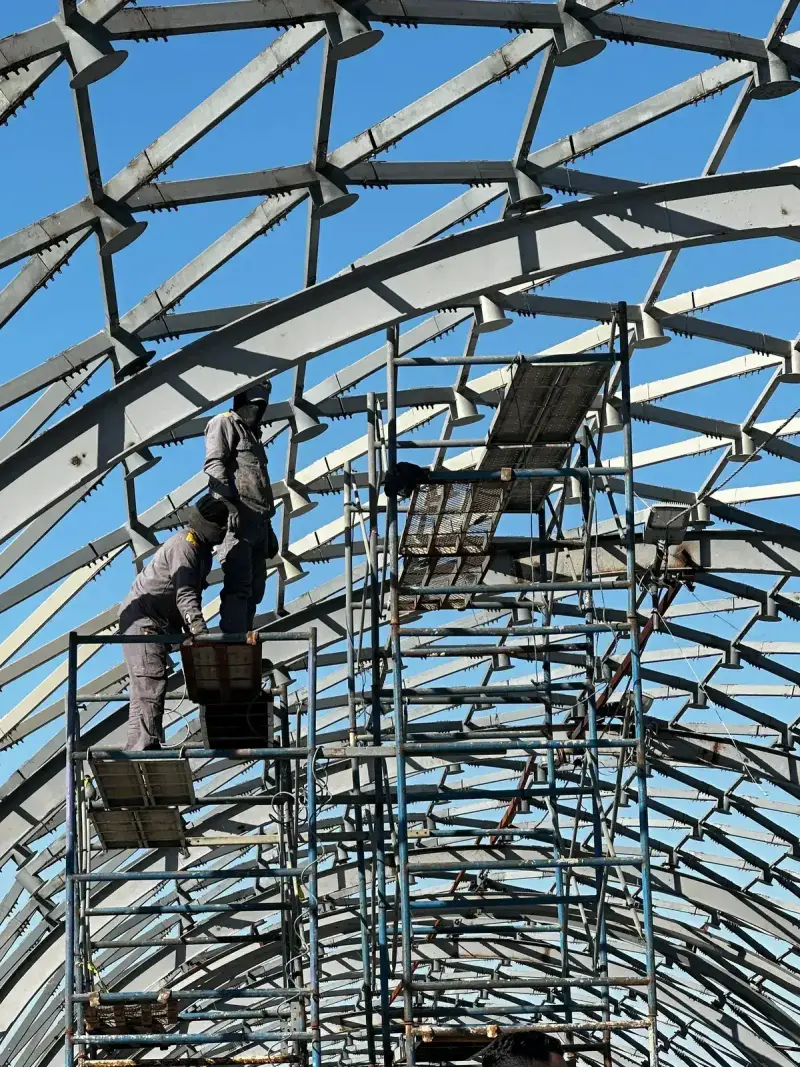 Workers on scaffolding inside a metal building frame against a clear blue sky